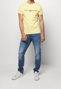 T-shirt en coton jaune avec impression de logo, associée à un jean en denim bleu et des baskets blanches. Le modèle se tient devant un fond gris clair.