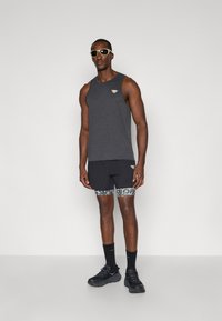 Μαύρο αθλητικό τοπ χωρίς μανίκια με λογότυπο, συνδυασμένο με μαύρες βερμούδες με διακοσμητική φάσα, μαύρες κάλτσες και μαύρα αθλητικά παπούτσια.