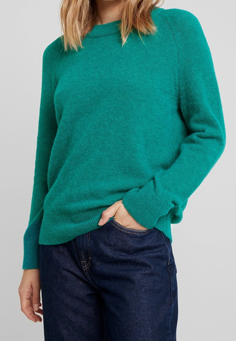 Türkiser, gestrickter Pullover mit Rundhalsausschnitt, langen Ärmeln und einer lockeren Passform, kombiniert mit dunkelblauen Jeans. Weiche Textur, keine sichtbaren Muster.