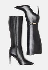 Bottes en cuir noir montantes jusqu'aux genoux, avec bouts pointus, talons stilettos, fermeture éclair latérale sur une botte, et sangle décorative avec boucle dorée sur l'autre.