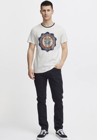 Hvit t-skjorte med en sirkulær grafikk i blått og oransje, svarte jeans og svarte joggesko med hvite detaljer. Korte ermer, rund hals.