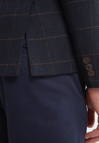 Jūrų laivyno houndstooth tipo blazeris su rudais persikėlimais, pagamintas iš tekstūruoto audinio. Turi šoninius ventiliacijos angas ir tris rudus mygtukus ant rankovės.