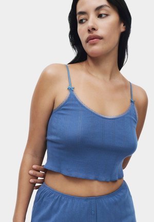 Kobieta o długich ciemnych włosach, ubrana w niebieski prążkowany crop top na cienkich ramiączkach oraz dopasowane niebieskie spodnie, patrząca w swoją prawą stronę.