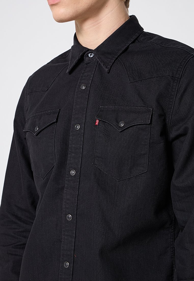 Camicia in denim nero con maniche lunghe, colletto a punta e chiusura con bottoni a scatto. Presenta due tasche sul petto e un dettaglio del logo rosso a contrasto.