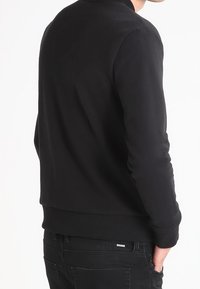 Svart sweatshirt med rund halsringning och ribbade ärmslut. Slät textur, figursydd design med en subtil logotyp på midjan.