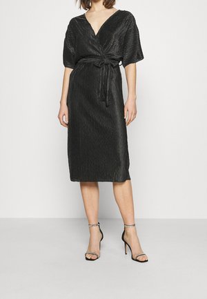 Kobieta ubrana w czarną, teksturowaną sukienkę kopertową z wiązaniem w talii i krótkimi rękawami, zestawioną z czarnymi sandałami na wysokim obcasie z paskami wokół kostek.