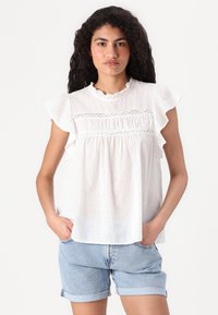 Blouse blanche à manches courtes avec des épaules à volants, présentant un motif de points texturés et des détails smockés au niveau de la poitrine. Assortie avec un short en denim.