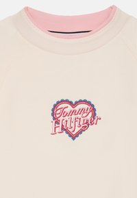 Jasno beżowy sweter z różowym ściągaczem i haftowanym logo w formie serca w kolorze czerwonym i niebieskim z napisem "Tommy Hilfiger".