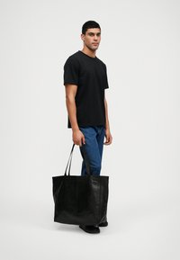 Άνδρας που φοράει μαύρη μπλούζα και μπλε τζιν κρατάει μια μεγάλη μαύρη δερμάτινη τσάντα ώμου σε μονόχρωμο φόντο.