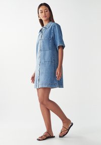 Lichtblauwe denim jurk met een kraag, korte mouwen, een knoopsluiting aan de voorkant en een zak aan de linkerkant.