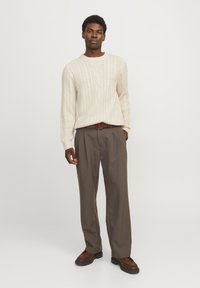 Cream cable-knit sweater featuring a round neckline and ribbed cuffs, paired with loose-fitting dark brown trousers and brown shoes.