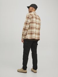 Karierte Flanellbluse in Beige- und Brauntönen, schwarze Cargo-Hosen und beigen Schuhe. Das Modell steht leicht abgewandt und trägt eine schwarze Mütze.