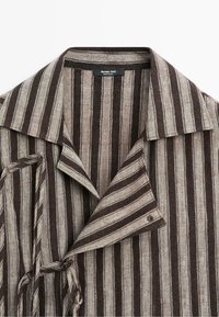 Chemise à col avec des rayures verticales marron et beige, montrant les boutons supérieurs et un détail de nœud en tissu sur un côté.