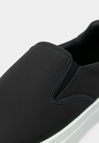 Chaussure noire à enfiler en matériau synthétique texturé avec des panneaux latéraux élastiques. Comprend une semelle en caoutchouc blanche et un design de bout lisse et arrondi.