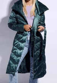 Abrigo acolchado de satén verde con un gran cuello, cintura ceñida con cinturón y acolchado en forma de diamante. Llevado sobre una blusa de color púrpura claro y pantalones de mezclilla.