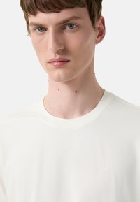Hvid t-shirt med korte ærmer lavet af glat stof, der har et subtilt præget "B" logo på brystet og en klassisk rund halsudskæring.