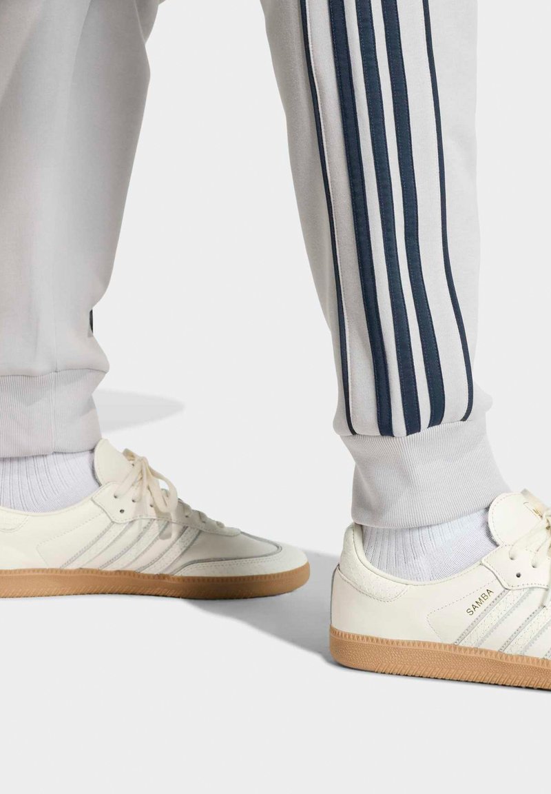 Jambes portant un pantalon de survêtement blanc à rayures bleu marine et des baskets blanches Adidas Samba avec semelles en gomme et des chaussettes blanches sur un fond clair.