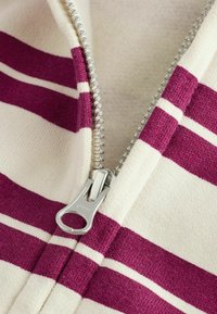 Vêtement zippé en tissu crème avec des rayures horizontales bordeaux, tricot texturé et détail de zip YKK argenté.
