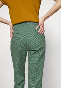 Personne portant un pantalon à carreaux verts et blancs et une chemise jaune moutarde, les mains posées sur les hanches, vue de dos.