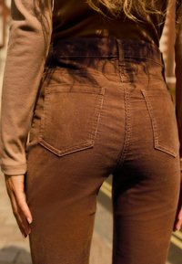 Pantalons en velours côtelé marron taille haute avec deux poches arrière, présentant des côtes verticales et une silhouette ajustée. Texture lisse, détails minimaux.