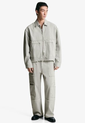 Giacca di jeans oversize grigia chiara con zip frontale e due tasche applicate, abbinata a pantaloni larghi coordinati con più tasche.