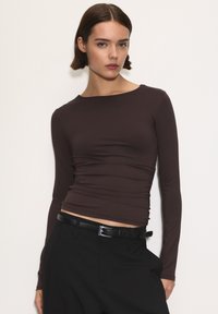 Brązowa bluzka z długim rękawem o dopasowanym kroju i marszczonym wykończeniu w talii, zestawiona z czarnymi spodniami o wysokim stanie i cienkim paskiem.