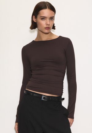 Smeđi dugosleeve top s uskim dizajnom i nabranom teksturom u struku, uparen s crnim hlačama visokog struka i tankim remenom.