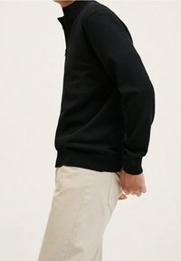 Suéter negro con cuello alto, mangas largas y puños acanalados. Combinado con pantalones de color beige claro. Diseño simple y ajustado.