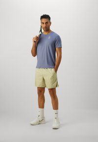 Lys lilla atletisk t-shirt med logo, parret med lys gule mønstrede shorts. Hvide sneakers og sokker, mens hun holder en tennisketsjer.