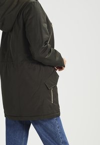 Olivově zelená parka se zipem uprostřed, kapucí a bočními kapsami, se mírně zúženým lemem. Hladká tkanina s matným povrchem.