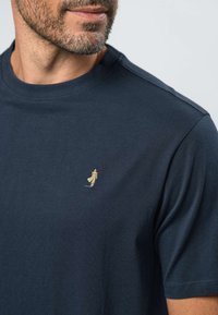 Mies, joka pukeutunut tummansiniseen pyöreäkauluksiseen T-paitaan, jossa on vasemmassa rinnassa pieni beige kirjailtu logo henkilöstä takki yllään ja hattu päässään.