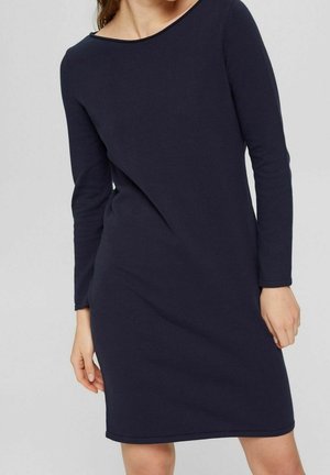 Femme portant une robe ajustée, manches longues, longueur genoux, bleu marine foncé avec un col rond simple, sur un fond clair uni.