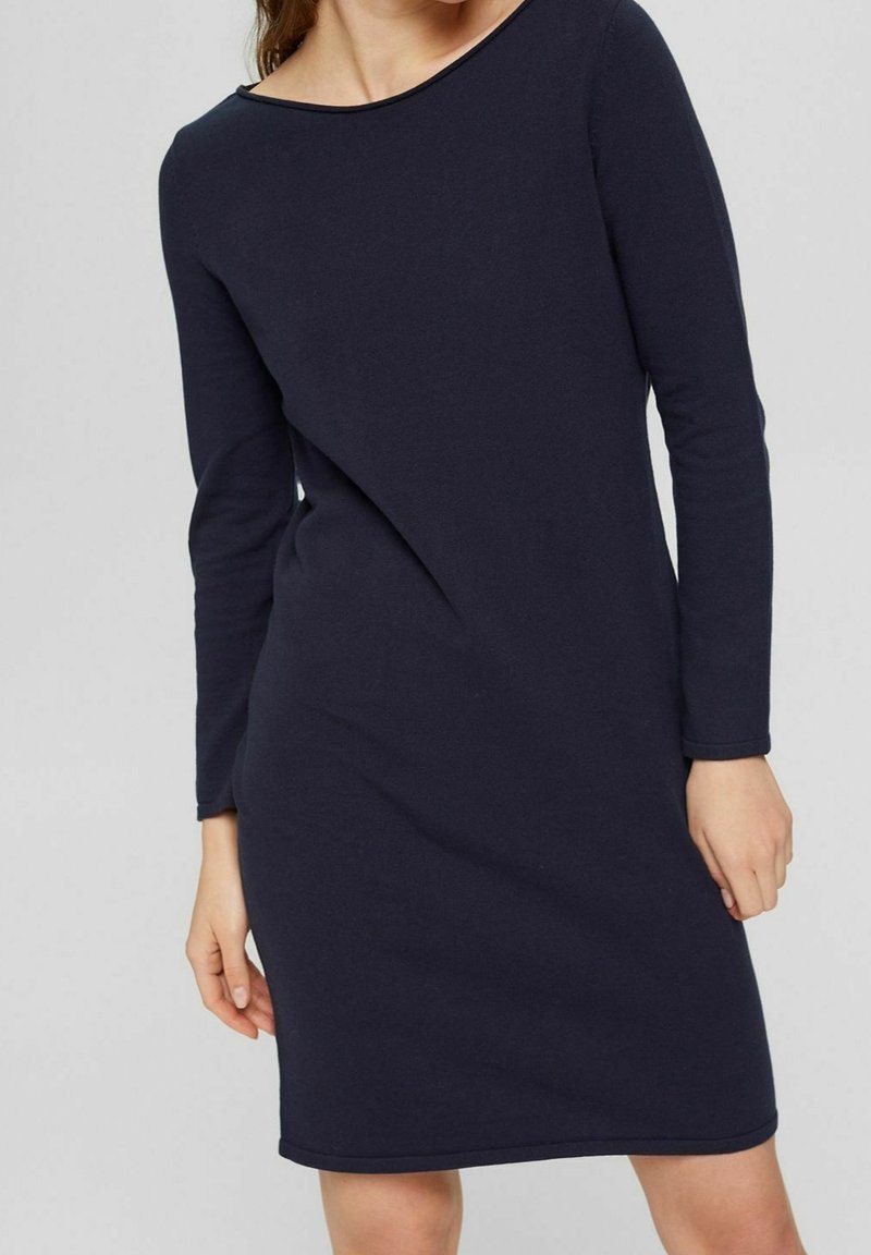 Femme portant une robe ajustée, manches longues, longueur genoux, bleu marine foncé avec un col rond simple, sur un fond clair uni.