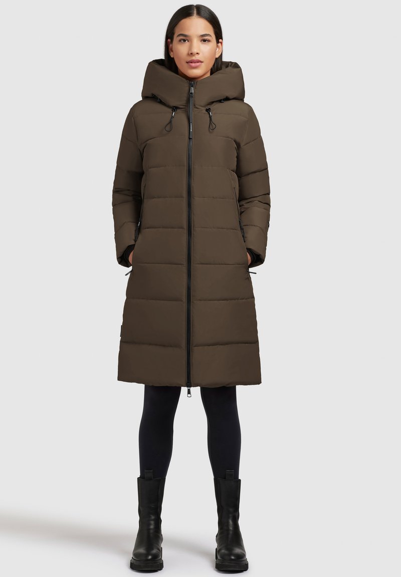 Frau in einem knielangen, braunen, gepolsterten Mantel mit hohem Kragen, schwarzen Leggings und schwarzen Stiefeletten, stehend vor einem einfarbigen, hellen Hintergrund.