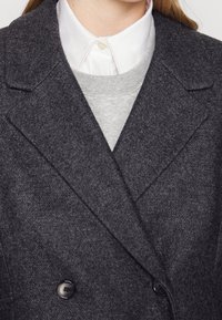 Abrigo gris de doble botonadura hecho de tejido texturizado, con solapas muesca y dos botones negros, superpuesto a un suéter gris y una camisa blanca.