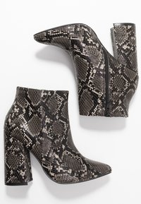 Stivali ankle con tacco alto, in pelle di serpente nera e grigia, punta affilata, tacco a blocco e zip laterale su sfondo bianco.