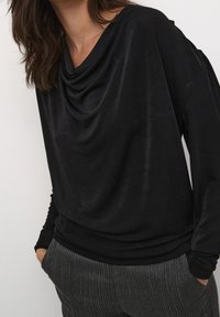 Svart draperad topp med en cowl-halsringning, mjukt tyg, långa ärmar och avslappnad passform, ihopparad med texturerade grårandiga byxor.