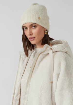 Kremowa dzianinowa czapka beanie z prążkowanym wzorem, ozdobiona małym złotym logo. Noszona z jasnym, fakturowanym płaszczem wierzchnim.