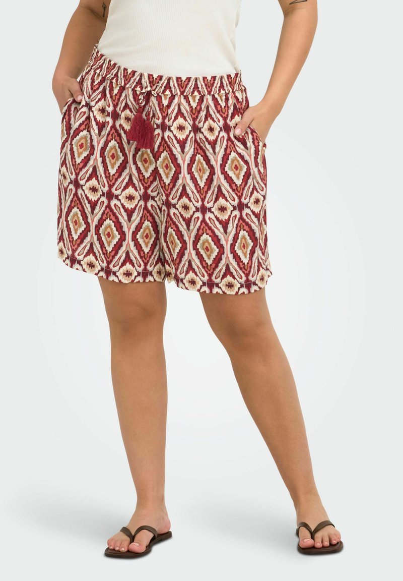 Femme portant un short rouge et beige à motif losanges avec un cordon à pompon noué, un haut blanc et des tongs noires, les mains dans les poches.
