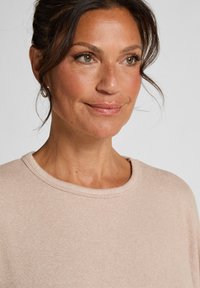 Lichtbeige trui met een ronde halslijn en een ribbelstructuur, gedragen door een vrouw met bruin haar in losse golven gestyled.