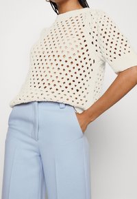 Sweter dzianinowy w kolorze off-white z fakturowanym, otwartym wzorem diamentowym i krótkimi rękawami, zestawiony z jasnoniebieskimi, dopasowanymi spodniami.