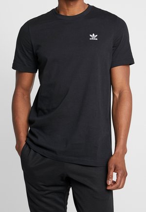 Mężczyzna ubrany w prostą czarną koszulkę Adidas z białym logo trefoil na piersi, stojący z jedną ręką w kieszeni na szarym tle.