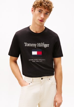 Jeune homme aux cheveux bouclés portant un t-shirt noir Tommy Hilfiger et un pantalon blanc, debout avec une main dans la poche devant un fond uni.