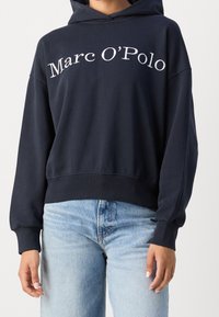 Sudadera azul marino hecha de tela suave, con un logotipo frontal "Marc O'Polo" en blanco, con un corte relajado y puños de canalé. Llevada con denim claro.