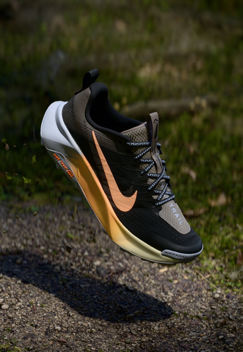 Zwarte en beige Nike trailhardloopschoen met oranje swoosh en witte zool, in de lucht boven een grindpad met groene bladeren op de achtergrond.