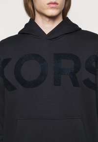 Sudadera negra con capucha que presenta un gran logo texturizado de "KORS" en azul marino. Incluye un bolsillo frontal y una textura de tela suave.