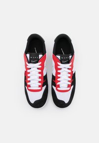Sportskor med en vit meshkropp, röda accenter på sidorna, en svart mockahæl och platta vita snörningar. Har en logotyp på tungan.