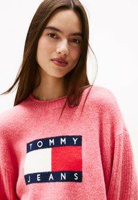 Junge Frau mit langen braunen Haaren, die einen rosa Pullover trägt, auf dem sich das Tommy Jeans-Logo in Weiß, Rot und Marineblau auf der Vorderseite befindet.