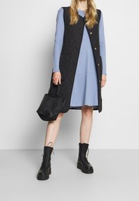 Svart vadderad väst med guldfärgad knappar, lagd över en ljusblå klänning. Svarta snörkängor och en liten svart toteväska fulländar looken.