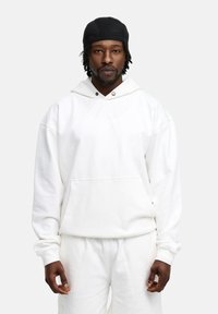 White hooded sweatshirt with a front pocket, loose fit, and ribbed cuffs. The model wears matching white pants and a black cap.
